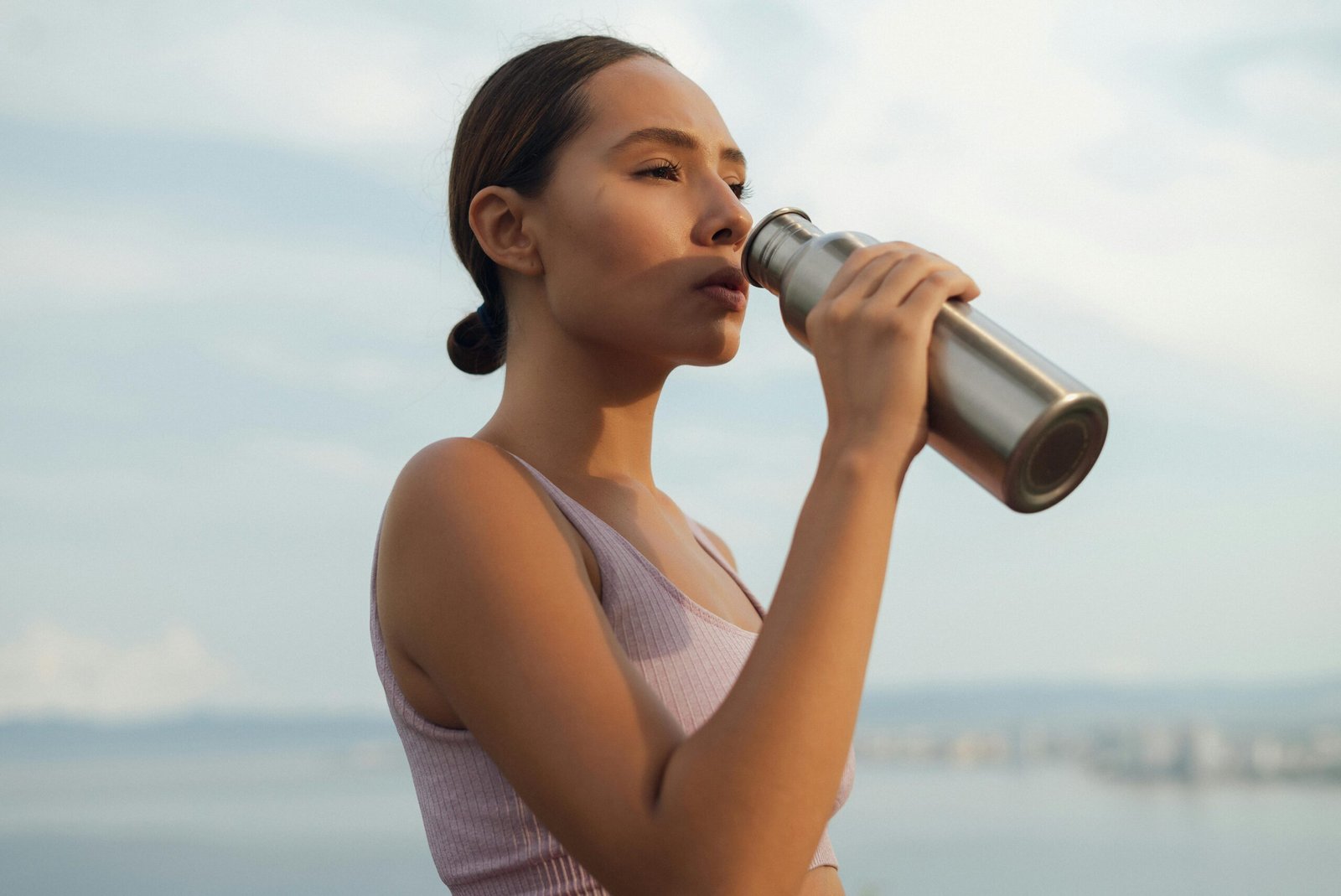 A woman drinks from a metal bottle on a sunny day by the beach, wearing a sports bra.